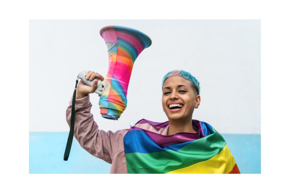 Young woman holding rainbow megaphone and wearing an LGBTQIA+ flag. Looking to come out and need guidance? LGBTQIA+ Affirmative Therapy in Evanston, IL can help you. Speak with an LGBTQIA+ affirming therapist in Illinois today to learn more about LGBTQ+ Affirming Therapy.