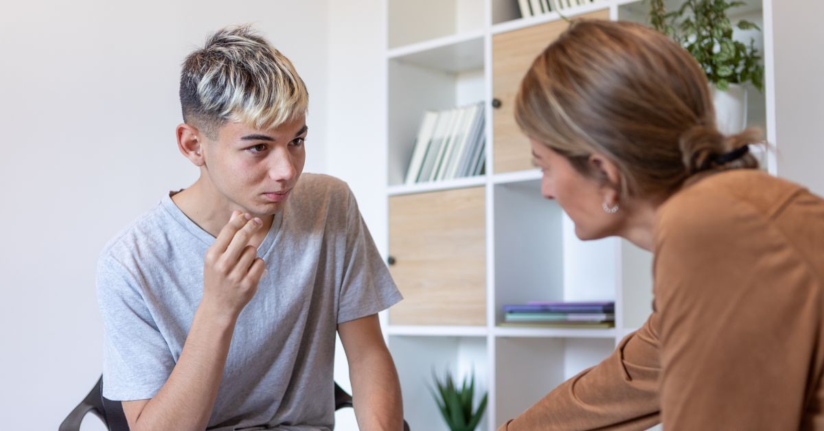 A teen boy sits in therapy with his therapist. Looking to see how a teen therapist can help your teen through Therapy for Teenagers? Learn more about it today to see how it helps teens!