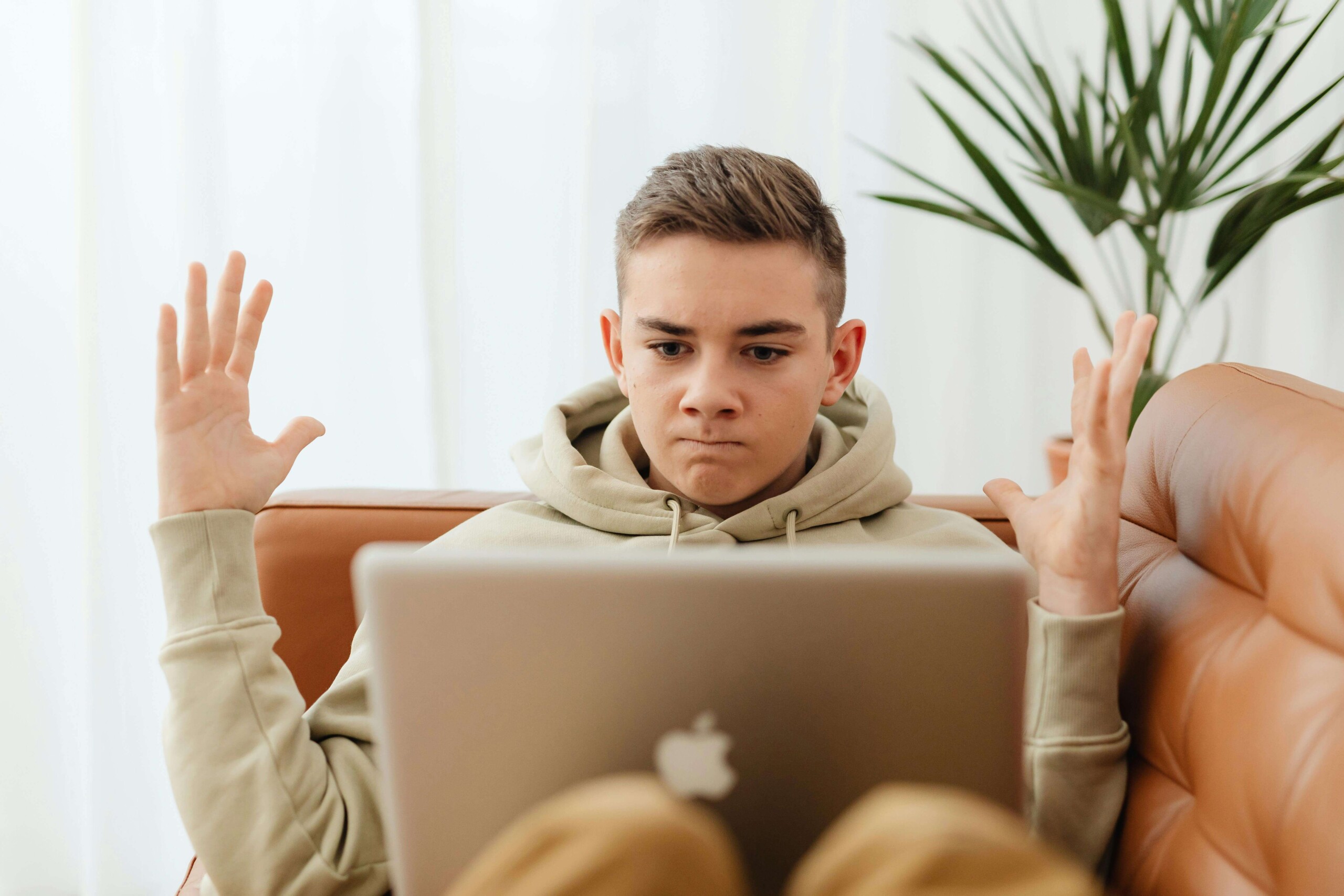 Frustrated teenage boy sitting on a couch with a laptop, raising his hands in confusion—capturing a moment that reflects the emotional challenges addressed through teen counseling in Evanston, IL and the importance of maternal mental health in Evanston, IL for parents supporting their teens.