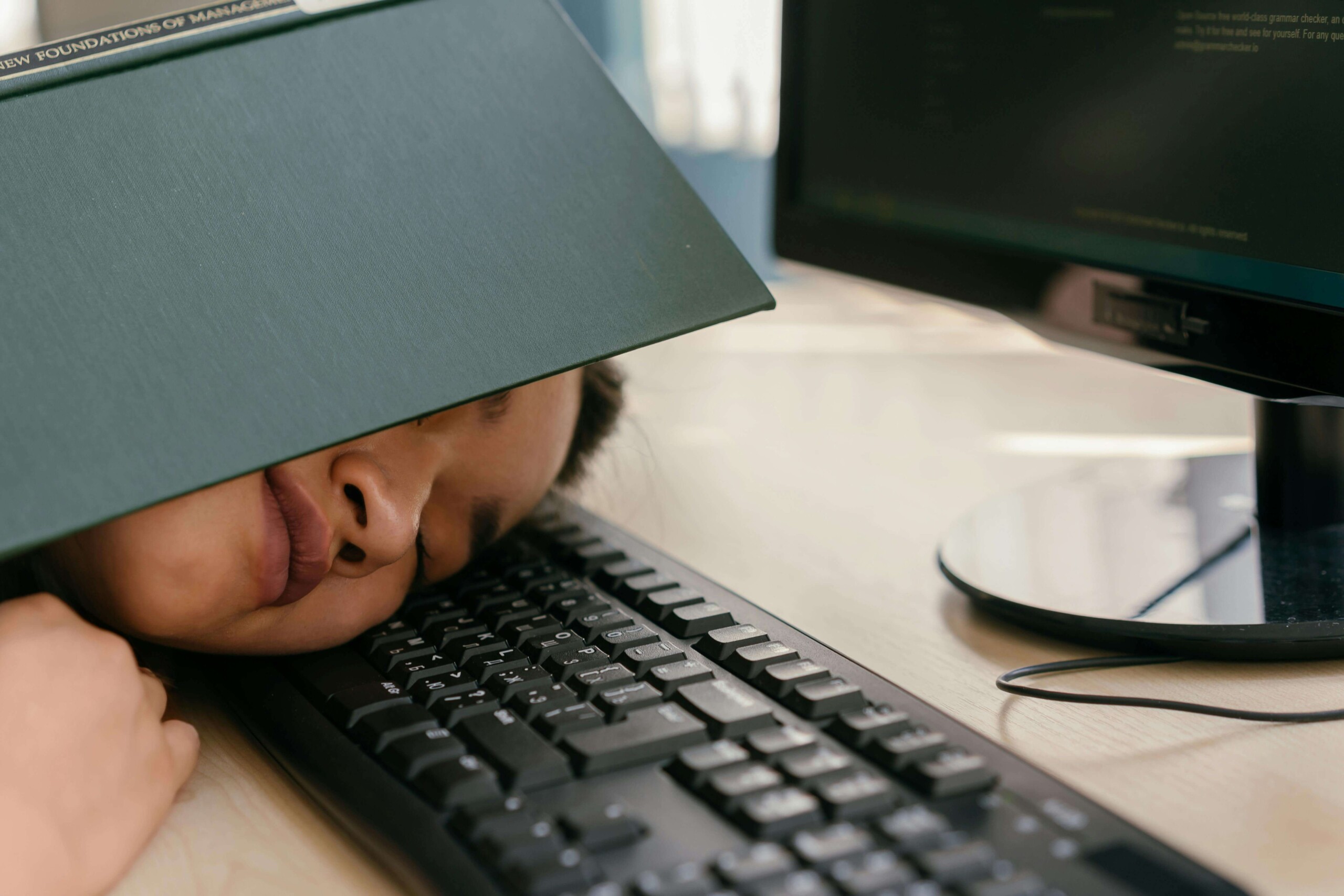 A college student asleep on a keyboard with a textbook covering their head, highlighting academic burnout and stress—representing the need for online therapy for college students in Chicago, IL and support during life transitions in Evanston, IL.