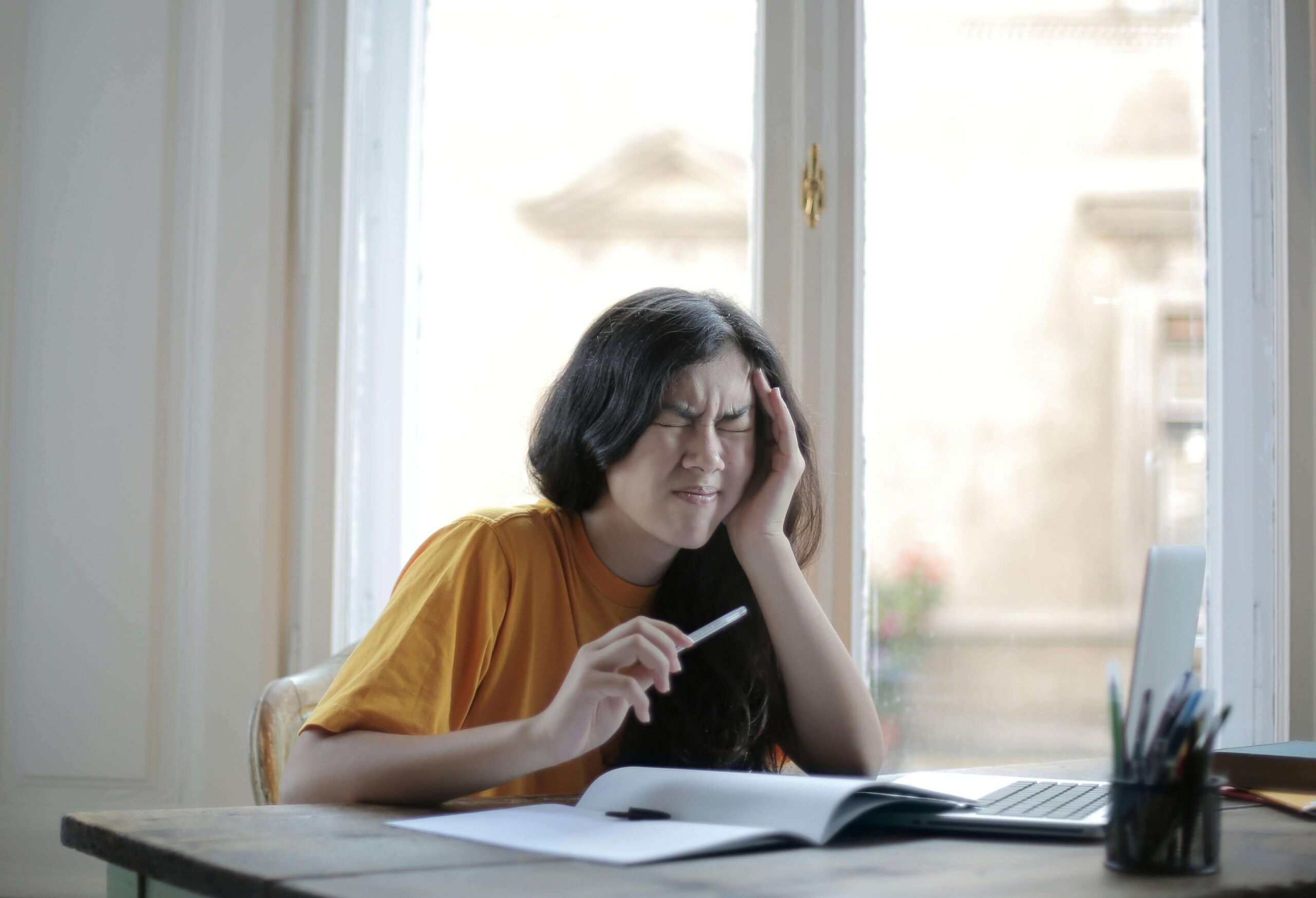 Young woman holding her head in pain while studying, representing chronic stress and cognitive overload—topics addressed in cognitive behavioral therapy in Chicago, IL by a young adult therapist in Illinois.