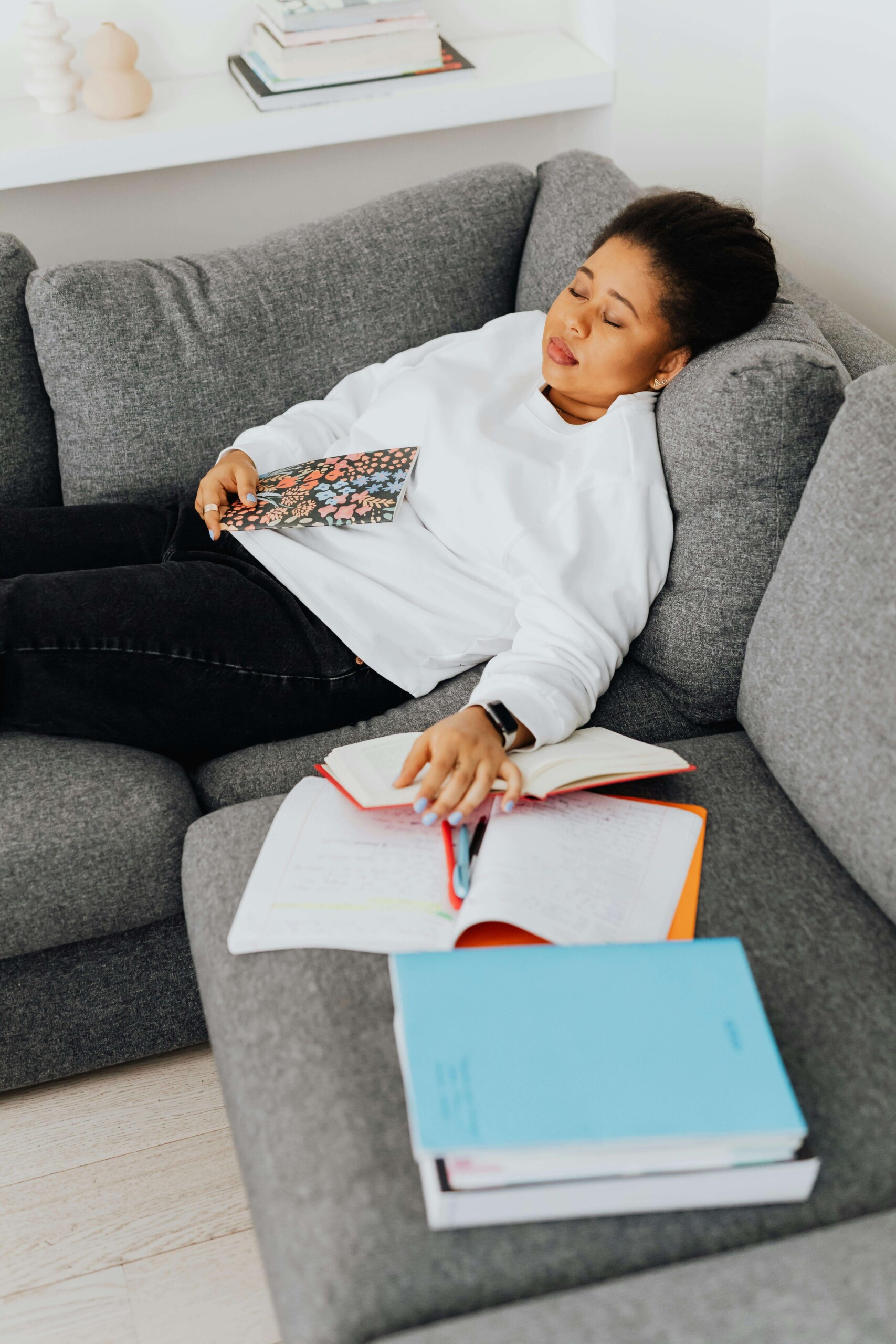 Exhausted from Assignments | young adult therapist in illinois | cognitive behavioral therapy in chicago, il | online therapy for college students in chicago, il | social anxiety in college evanston | Rogers Park, IL | Streeterville, IL | Northwestern University, IL High-angle view of a young woman resting on a gray sofa surrounded by books and notebooks, symbolizing academic stress and social anxiety in college Evanston. Ideal for a blog on how a young adult therapist in Illinois can help students manage pressure and anxiety.