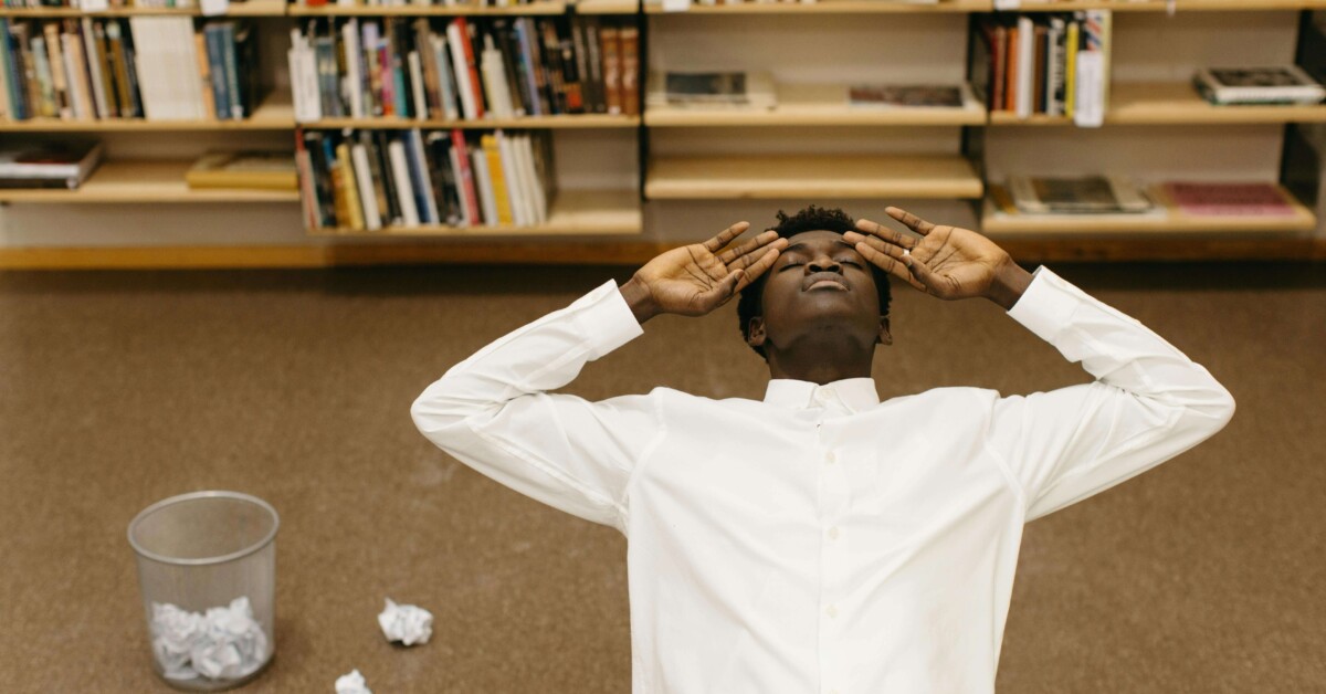 Overhead view of a college student lying on the floor in a library, surrounded by books and crumpled papers, symbolizing academic stress. Perfect visual for a blog on how cognitive behavioral therapy in Chicago, IL, and therapists for college students in Chicago, IL, can help manage academic pressure.