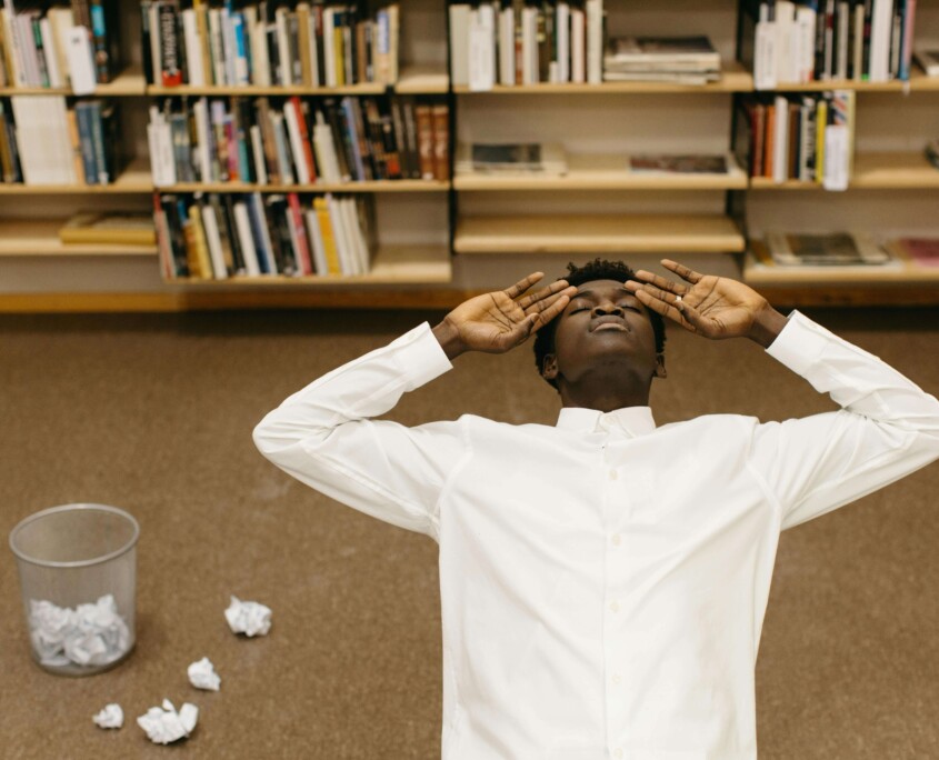 Overhead view of a college student lying on the floor in a library, surrounded by books and crumpled papers, symbolizing academic stress. Perfect visual for a blog on how cognitive behavioral therapy in Chicago, IL, and therapists for college students in Chicago, IL, can help manage academic pressure.