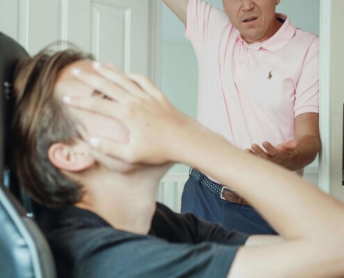 A young man sits in a chair, covering his face, while an older man stands in a doorway, gesturing with concern. Cognitive behavioral therapy in Chicago, IL, and a young adult therapist in Illinois can help families navigate holiday conflicts and improve communication.