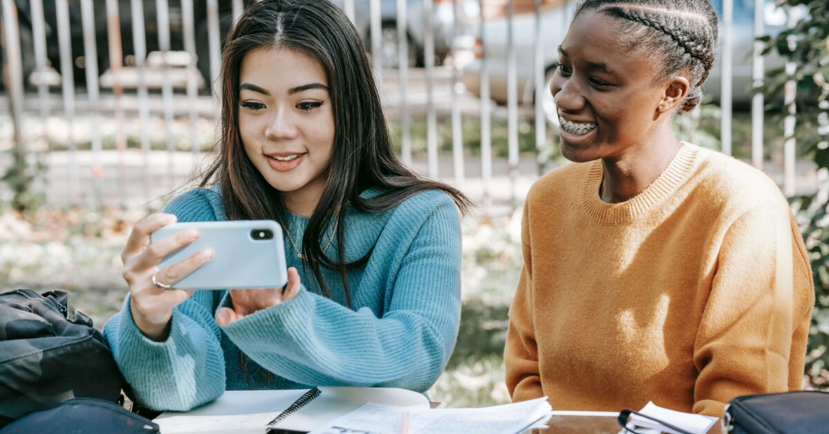 Two teen girls looking at a phone together while studying outdoors, illustrating the constant pull of social media and the pressure to perform happiness—challenges addressed through therapy for teens in Chicago, IL and cognitive behavioral therapy in Chicago, IL.