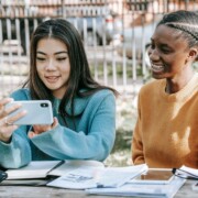 Two teen girls looking at a phone together while studying outdoors, illustrating the constant pull of social media and the pressure to perform happiness—challenges addressed through therapy for teens in Chicago, IL and cognitive behavioral therapy in Chicago, IL.
