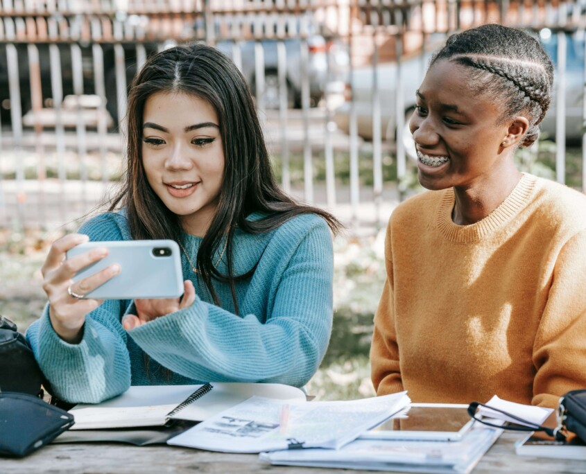 Two teen girls looking at a phone together while studying outdoors, illustrating the constant pull of social media and the pressure to perform happiness—challenges addressed through therapy for teens in Chicago, IL and cognitive behavioral therapy in Chicago, IL.