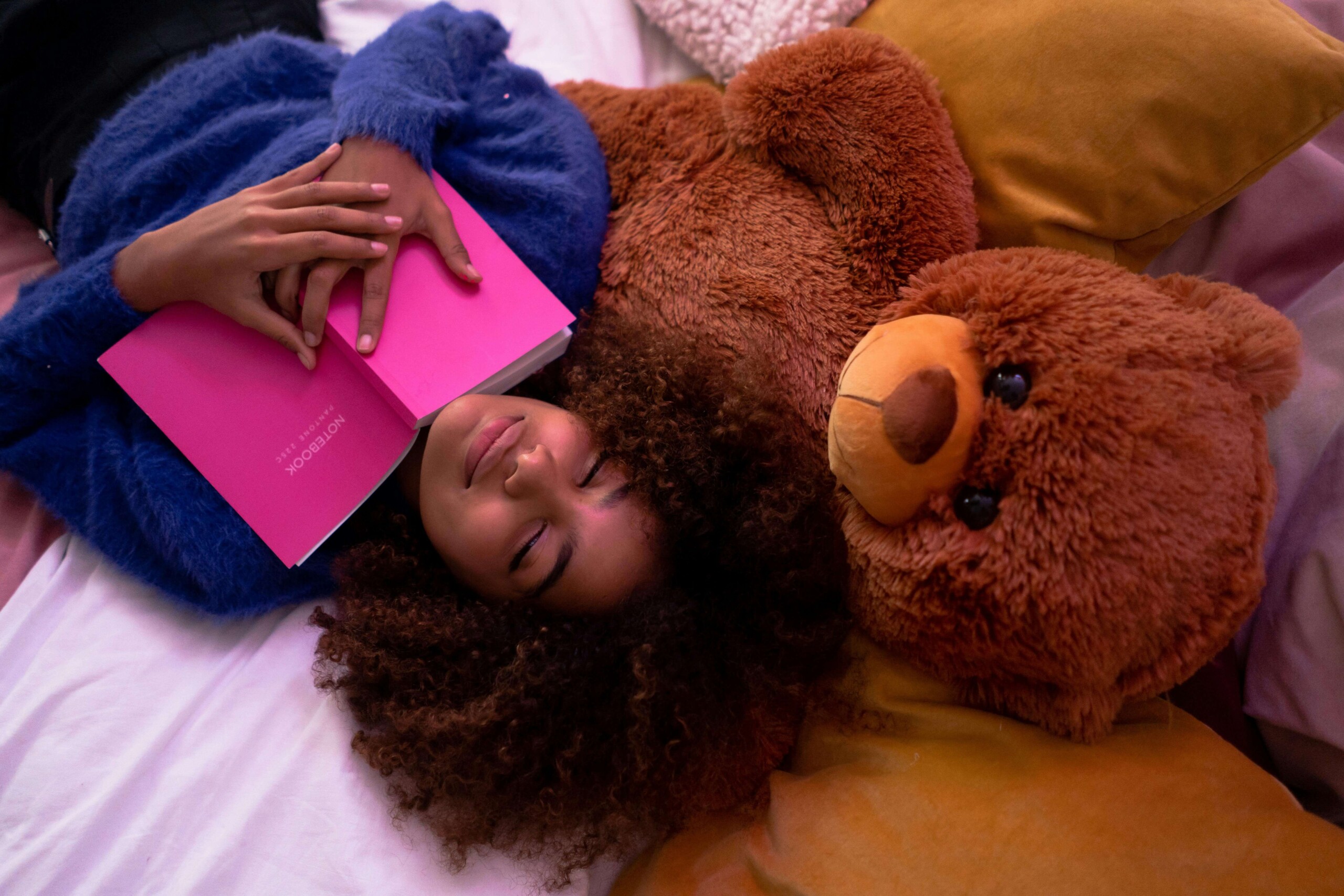 A teen girl lying on her bed with a book covering her face, surrounded by stuffed animals, capturing the exhaustion of maintaining a happy facade—feelings that can be processed through online therapy for teens in Chicago and online college counseling in Chicago, IL.