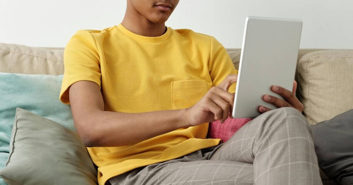 A teen sitting on a couch using a tablet at home, illustrating the accessibility of working with an online therapist for teens in Evanston, IL through online therapy for teenagers in Evanston, IL.