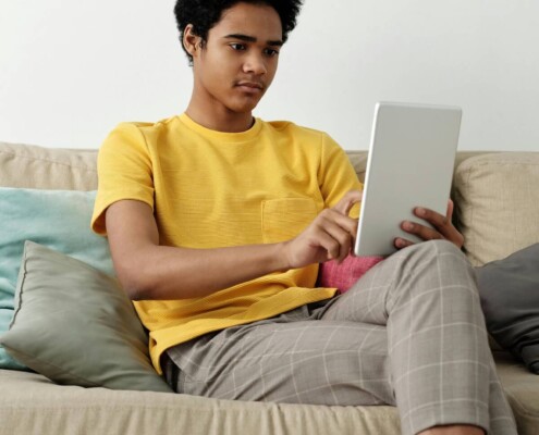 A teen sitting on a couch using a tablet at home, illustrating the accessibility of working with an online therapist for teens in Evanston, IL through online therapy for teenagers in Evanston, IL.