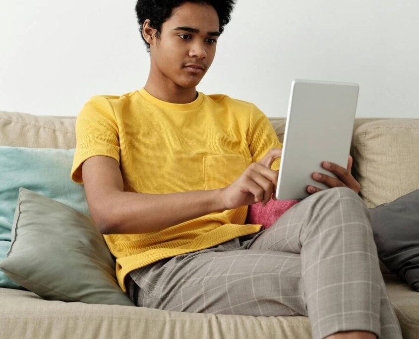 A teen sitting on a couch using a tablet at home, illustrating the accessibility of working with an online therapist for teens in Evanston, IL through online therapy for teenagers in Evanston, IL.