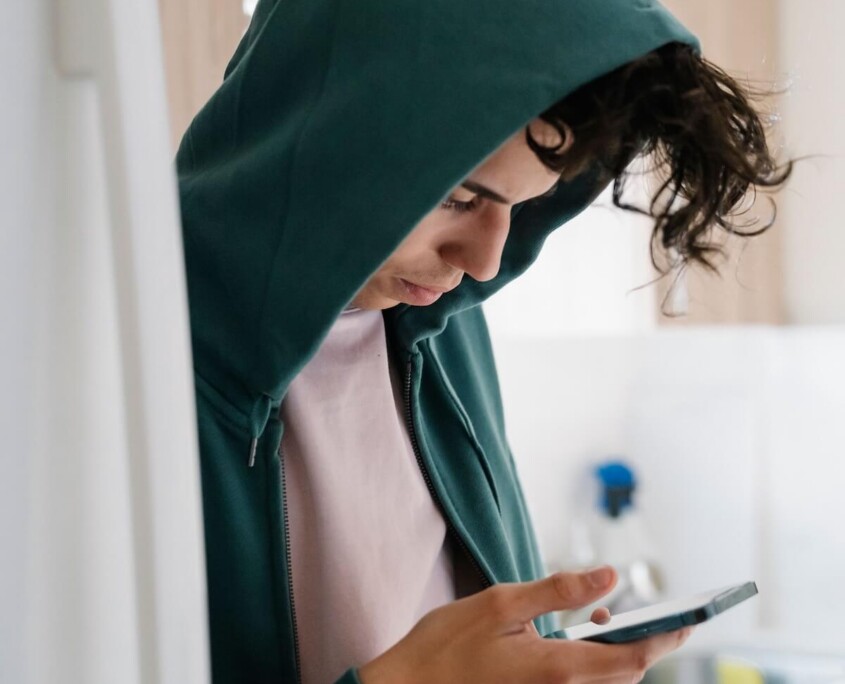 A teen boy in a hoodie looking down at his phone in the kitchen, representing the constant exposure to online content that can shape his identity and may call for support from an Evanston teen therapist or teenage intervention in Evanston, IL.