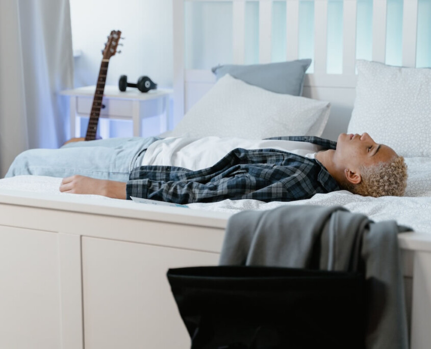 A teen boy lying on his bed with his backpack nearby, representing the emotional exhaustion of school avoidance that can be addressed through teen therapy in Evanston, IL with support from an Evanston teen therapist.